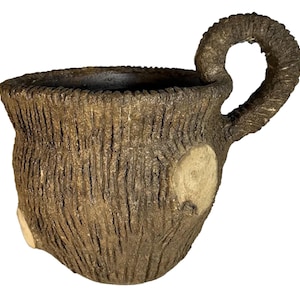 Puede incluir: Una taza de cerámica marrón con textura de corteza de árbol y un asa gruesa y retorcida. La taza tiene dos acentos circulares blancos en el lateral.