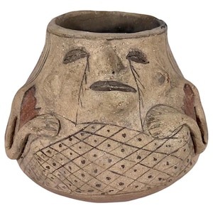 May include: A tan ceramic vessel featuring a sculpted face with closed eyes and a mouth. The lower portion displays a cross-hatched grid pattern with small dots in each section. Two sculpted arms are positioned on the sides of the pot.