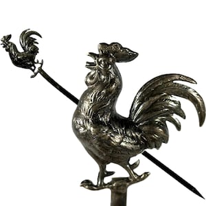 May include: A silver-toned metal rooster weathervane with a detailed design. The rooster is depicted crowing, with a smaller version of the rooster on a rod. The weathervane is designed to be mounted on a roof or structure to indicate wind direction.