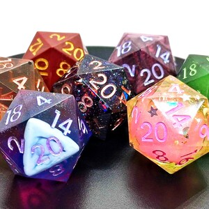May include: A collection of polyhedral dice in various colors, including red, green, purple, and gold. Each die features visible numbers and intricate designs, perfect for tabletop gaming. The dice are arranged on a dark surface.