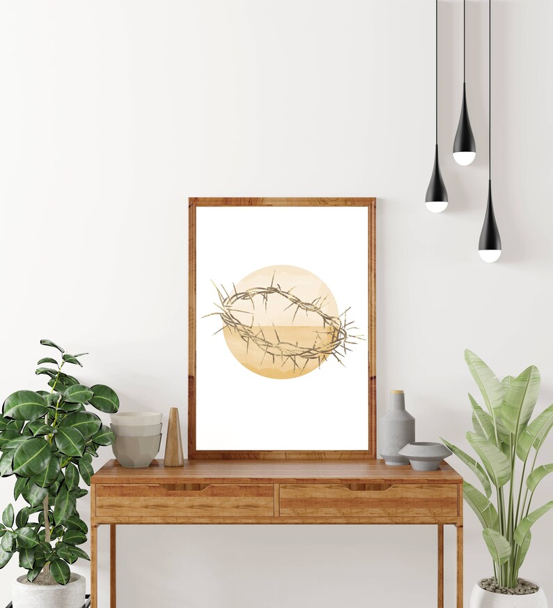May include: A framed print of a watercolor illustration of a crown of thorns on a yellow circle. The print is hanging on a white wall above a wooden table.