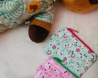 May include: Two small card holders with zipper closures. One is green with a white and pink cartoon design. The other is pink with a white and green cartoon design.