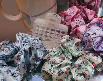 May include: A collection of colourful hair scrunchies with various patterns, including a blue and white scrunchie with a floral design, a green and white scrunchie with a skull and crossbones design, and a purple and white scrunchie with a star design.