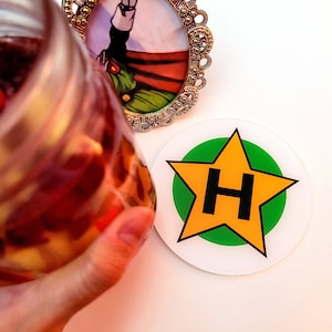 Op de afbeelding: Een witte sticker met een groene cirkel en een gele ster met een zwarte "H" in het midden. De ster heeft zes punten.