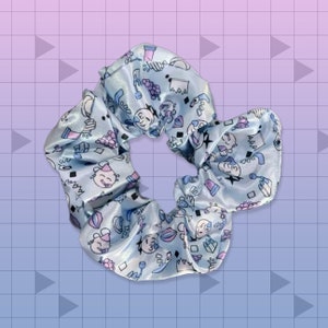 Puede incluir: Un scrunchie azul y blanco con un patrón de varios diseños gráficos pequeños. El scrunchie está hecho de tela de satén y tiene una apariencia arrugada y abullonada.