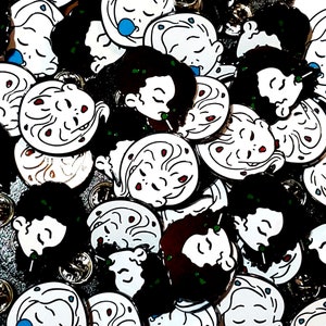 Puede incluir: Una colección de pines de esmalte con una variedad de diseños, que incluyen una ilustración en blanco y negro de una cara con ojos rojos y una ilustración en blanco y negro de una cara con un ojo azul. Los pines son todos redondos y tienen un respaldo plateado.