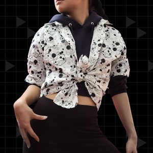 May include: A white button-down shirt with a black geometric pattern. The shirt is tied at the waist and worn over a black hooded sweatshirt.