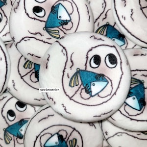 May include: A collection of round, plush, off-white fabric discs featuring a printed illustration of a wide-eyed creature holding a teal, triangular-shaped object with yellow fins. The text 'pecknorder' is printed in black on the central disc.