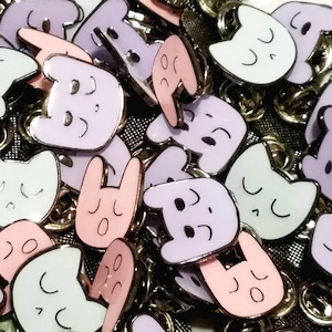 May include: A collection of enamel pins featuring various cartoon characters with different expressions. The pins are in various colors, including pink, purple, white, and light blue. The pins are all in the shape of a cartoon animal or creature.