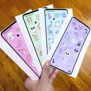 Può includere: Cinque custodie per telefono diverse con vari design e adesivi colorati. Ogni custodia ha il testo "I ECK N ORDER" stampato su di essa. Le custodie presentano una varietà di design, tra cui cuori, stelle, teschi e altre grafiche divertenti.