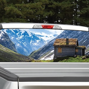Può includere: Un adesivo per finestrino di camion che presenta un paesaggio montano con un piccolo cottage di legno in primo piano. Le montagne sono coperte di neve e il cielo è blu.