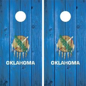 Può includere: Due tavole di cornhole in legno blu con cerchi bianchi e la bandiera dello stato dell'Oklahoma dipinta su ogni tavola. La bandiera presenta uno scudo dorato con una croce rossa e un ramo verde, circondato da piume bianche.