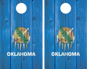 Puede incluir: Dos tableros de cornhole de madera azul con círculos blancos y la bandera del estado de Oklahoma pintada en cada tablero. La bandera presenta un escudo dorado con una cruz roja y una rama verde, rodeado de plumas blancas.