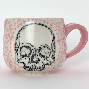 May include: A ceramic mug with a glossy white finish featuring a black ink illustration of a human skull on the front. The mug is decorated with a pink lace-like pattern surrounding the skull and has a solid pink handle on the right side.