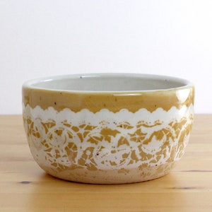Può includere: Una ciotola in ceramica con una glassa bianca e gialla. La ciotola ha un bordo smerlato e un design a pizzo.