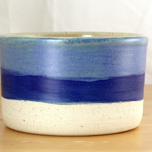 Pode incluir: Uma tigela de cerâmica branca com um esmalte azul e azul claro. A tigela tem uma textura salpicada e uma forma arredondada.