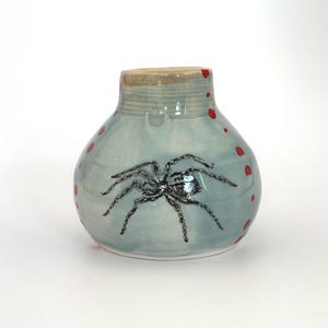 May include: A small, round, ceramic vase with a light blue glaze. A black spider is painted on the front, with red dots scattered around the vase. The rim is a tan color.