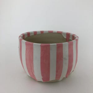 Puede incluir: Una maceta de cerámica blanca con rayas verticales rosas. La maceta tiene una forma redondeada y un borde ligeramente irregular.