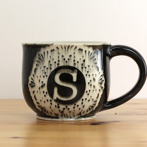 May include: Black and white ceramic mug with a white 'S' in a circle design. The mug has a textured, raised design around the circle.