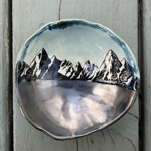 May include: A handmade ceramic bowl with a mountain range design. The bowl features a silver-colored base and a light blue upper section with a black and white mountain illustration. The rim is a darker blue. The bowl is irregularly shaped.