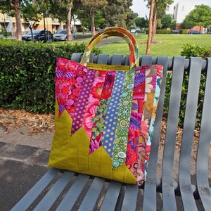 May include: A green patchwork tote bag with colourful fabric strips and floral patterns. The bag has two handles and is sitting on a metal bench.