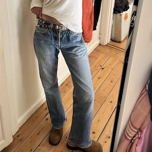May include: Faded blue denim jeans with a button fly and a worn appearance. The jeans are paired with a brown leather belt and brown shoes. The jeans have a classic, straight-leg fit.