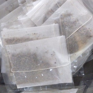 May include: A pile of individual tea bags, each containing loose tea leaves, are stacked on top of each other. The tea bags are made of white paper and are sealed with a string.