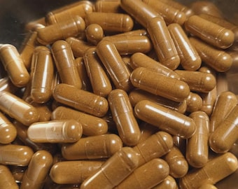 May include: A large collection of cylindrical capsules filled with a fine, light brown powder. Each capsule features a smooth, semi-translucent shell that reveals the granular texture of the contents inside, piled together in a metallic container.