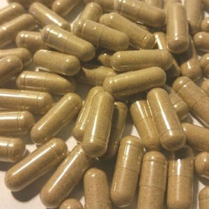 May include: A close-up shot of numerous brown capsules. The capsules are cylindrical with rounded ends and appear to be filled with a light brown powder. The capsules are scattered across a white surface.