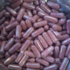 May include: A close-up image of a large quantity of dark purple capsules in a metal bowl. The capsules are translucent and show the dark purple powder inside.