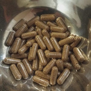 May include: A collection of numerous cylindrical capsules filled with a granular brown powder. The capsules are translucent, revealing the textured contents inside, and are gathered together in a polished, reflective metallic bowl.