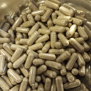 May include: A close-up view of a large quantity of brown capsules in a silver bowl. The capsules are filled with a light brown powder.