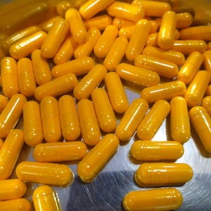 May include: A close-up shot of numerous yellow capsules, likely supplements or medications, filling a metallic bowl. The capsules are uniformly colored and cylindrical, suggesting a consistent product.