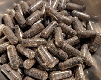 May include: A close-up of a metal bowl filled with a large quantity of dark gray capsules. The capsules are arranged in a random pattern, with some overlapping.