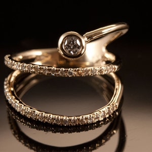 May include: A gold ring with a single diamond set in a bezel setting. The ring is designed with two bands, one of which is a simple band and the other is a curved band with a diamond set in the center.