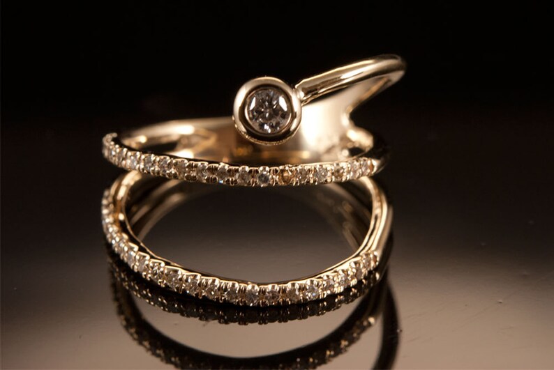 May include: A gold ring with a single diamond set in a bezel setting. The ring is designed with two bands, one of which is a simple band and the other is a curved band with a diamond set in the center.