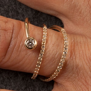 May include: A gold ring with three bands of pave diamonds and a single round diamond set in a bezel setting. The ring is designed to wrap around the finger.