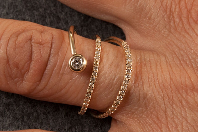 May include: A gold ring with three bands of pave diamonds and a single round diamond set in a bezel setting. The ring is designed to wrap around the finger.