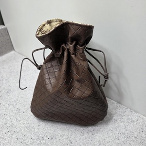 May include: A brown leather drawstring pouch with a woven pattern. The pouch is gathered at the top with a drawstring closure. The interior lining has a cream and brown pattern.