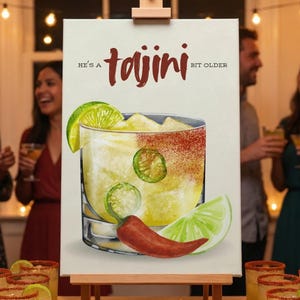 May include: A canvas print on an easel displays a watercolor illustration of a cocktail with lime, jalapeño slices, and a chili pepper. The text reads "He's a Tajini bit older". The image is set at a party with people holding drinks.