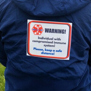 May include: A navy blue jacket with a white and red warning sign. The sign reads "WARNING! Individual with compromised immune system! Please, keep a safe distance!" in black and blue text.