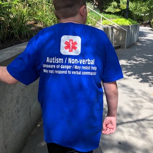 Könnte beinhalten: Ein königsblaues T-Shirt mit weißem Text und einem roten medizinischen Symbol. Der Text lautet "Autismus / Non-verbal" und enthält Sicherheitsinformationen. Das T-Shirt wird von einer Person getragen, die draußen geht.