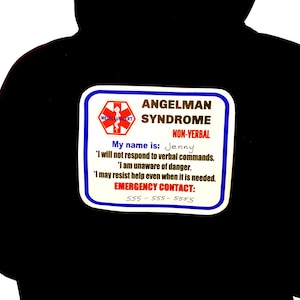 Puede incluir: Una sudadera con capucha negra con un parche de alerta médica blanco y azul. El parche dice "Síndrome de Angelman No Verbal" e incluye el nombre "Jenny" y la información de contacto de emergencia. El símbolo de alerta médica es visible.
