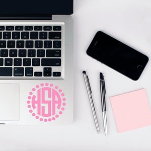May include: A silver laptop computer with a pink monogram sticker on the trackpad. The sticker features the letters "HSA" in a circle with pink dots around the edge. The laptop is on a white surface with a black smartphone, two silver pens, and a pink sticky note.