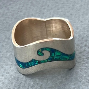 May include: Silver-toned ring with a wave design. The wave is inlaid with a mosaic of teal and green stones. The ring has a wide band and a smooth, polished finish, suitable for everyday wear.