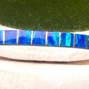 May include: A silver bracelet with a row of vibrant blue opal stones. The stones are rectangular and set in a silver band. The bracelet has a decorative design on the sides and is displayed on a white surface.