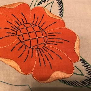 May include: A close-up of a hand-stitched orange flower with a black center and a brown outline. The flower is embroidered on a beige fabric.