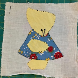 May include: A patchwork design on fabric, depicting a figure with a yellow head and three yellow body sections. The figure's dress is blue with red roses and yellow flowers. The background is white fabric.