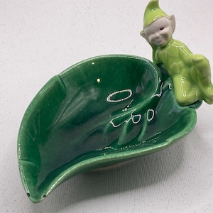 Puede incluir: Un plato de cerámica en forma de hoja verde brillante, con una figura de elfo reclinado en verde a juego. El elfo tiene una cara blanca y un gorro verde puntiagudo. Es un objeto decorativo.