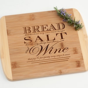 May include: A wooden cutting board with a light brown finish and a natural wood grain. The board has a rounded edge and is engraved with the text "BREAD that this house may never know hunger SALT that life may always have flavor and WINE that joy & prosperity may reign forever".